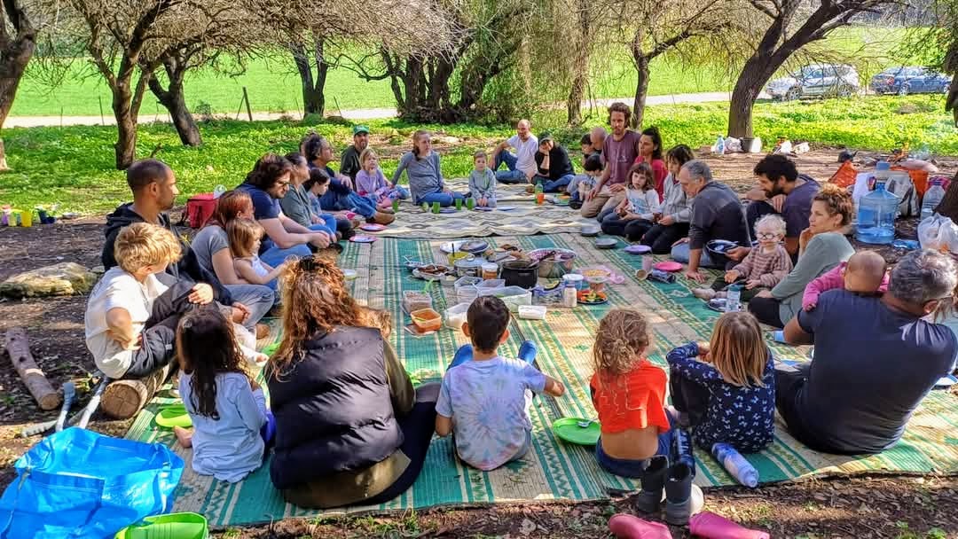משפחות יושבות במעגל על מחצלות ביער סביב ארוחה משותפת
